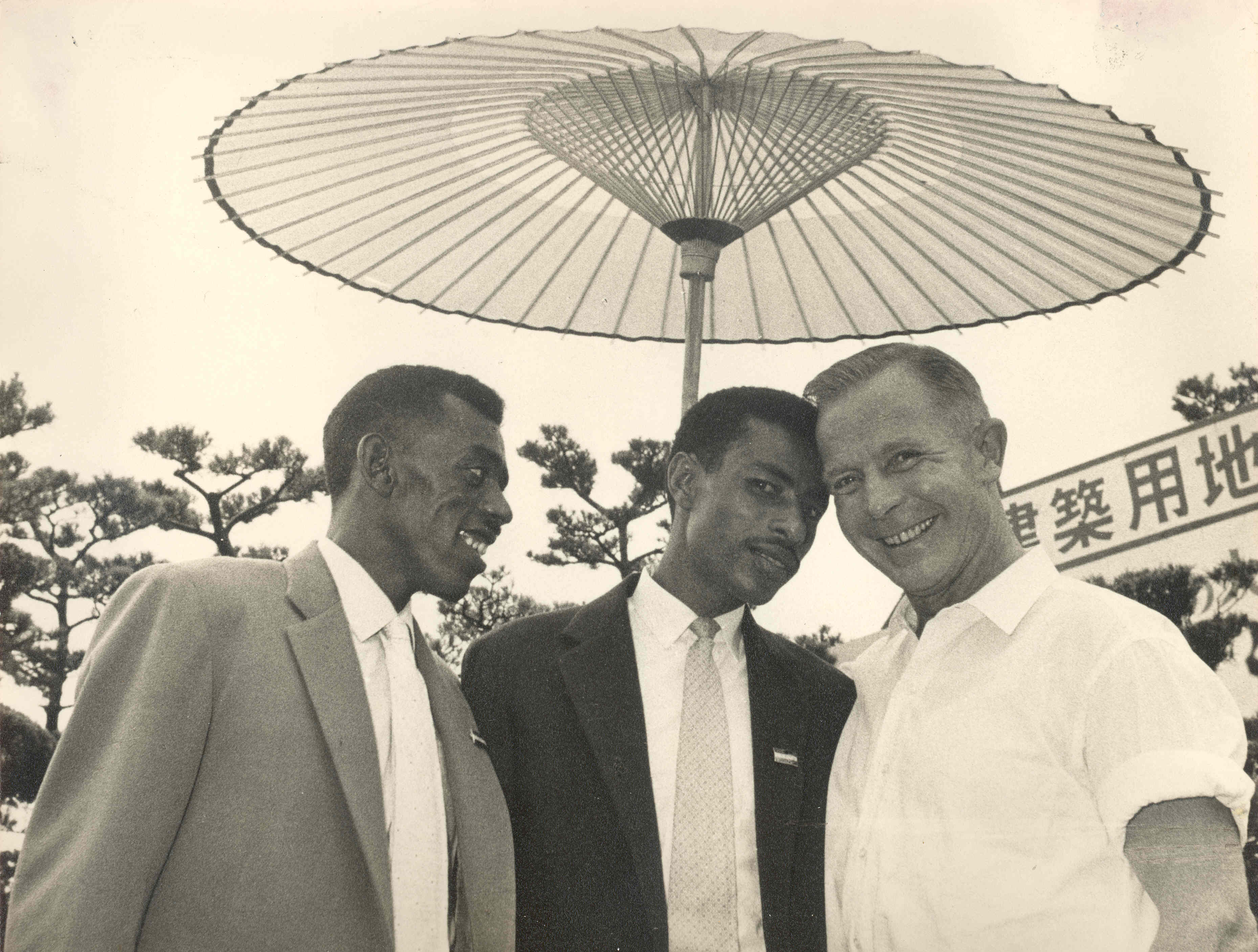 Three men beneath a Japanese paper parasol: a man in a light jacket, Abebe Bikila in a dark suit, and Onni Niskanen in a white shirt.