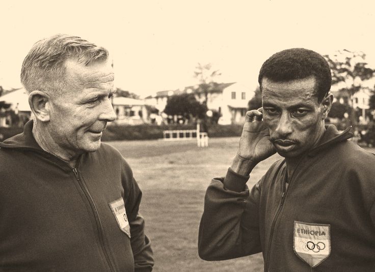 Onni Niskanen and Abebe Bikila in Ethiopia Olympic tracksuits; Niskanen looks toward Abebe, who rests his head on his hand.