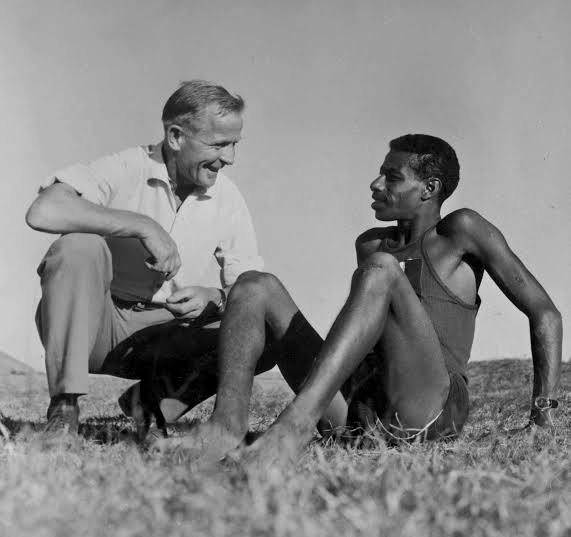 Onni Niskanen crouching and smiling beside Abebe Bikila, who sits barefoot on the grass in a running vest.