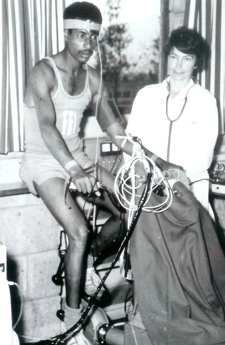 Abebe Bikila on a stationary bicycle wired to monitoring equipment, beside a woman in a white coat with a stethoscope.
