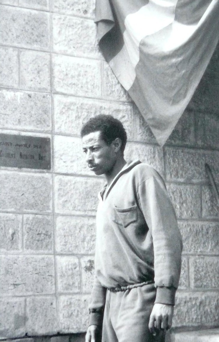 Abebe Bikila in a training top standing before a stone wall beneath a draped flag.