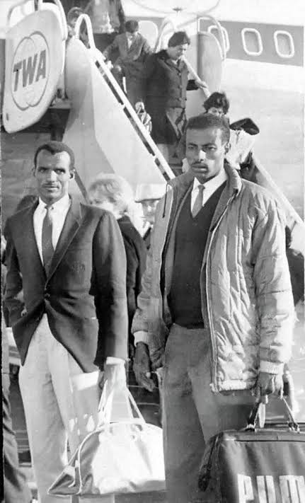 Abebe Bikila and a fellow traveller walk away from a TWA aircraft carrying their bags.