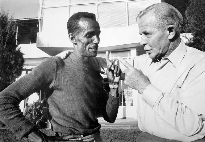 Abebe Bikila and Onni Niskanen in conversation, each raising a finger as if counting or making a point.